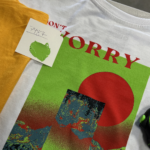A 100 Garment-Dyed Heavyweight Shirt - Text-Based Artwork with Vector Designs - Maximum of 2 Ink Colors - Soft Hand Screen Printing, with a colorful graphic and the text "DON'T WORRY," next to a yellow shirt and a green color sample swatch on a desk.